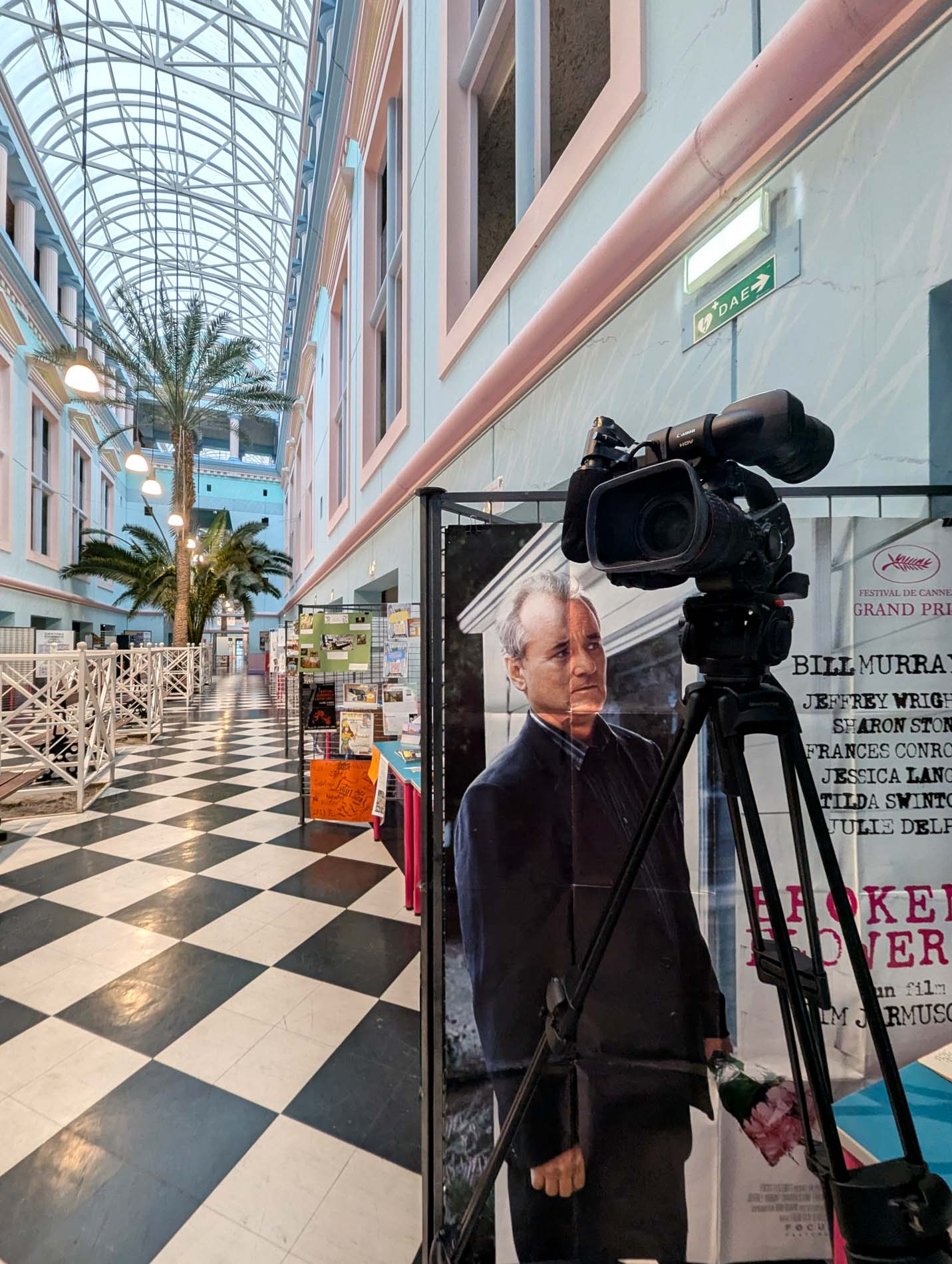 La rue intérieure : sol en damier, palmiers et verrière en voûte ; au premier plan, une caméra sur trépied devant une affiche de film